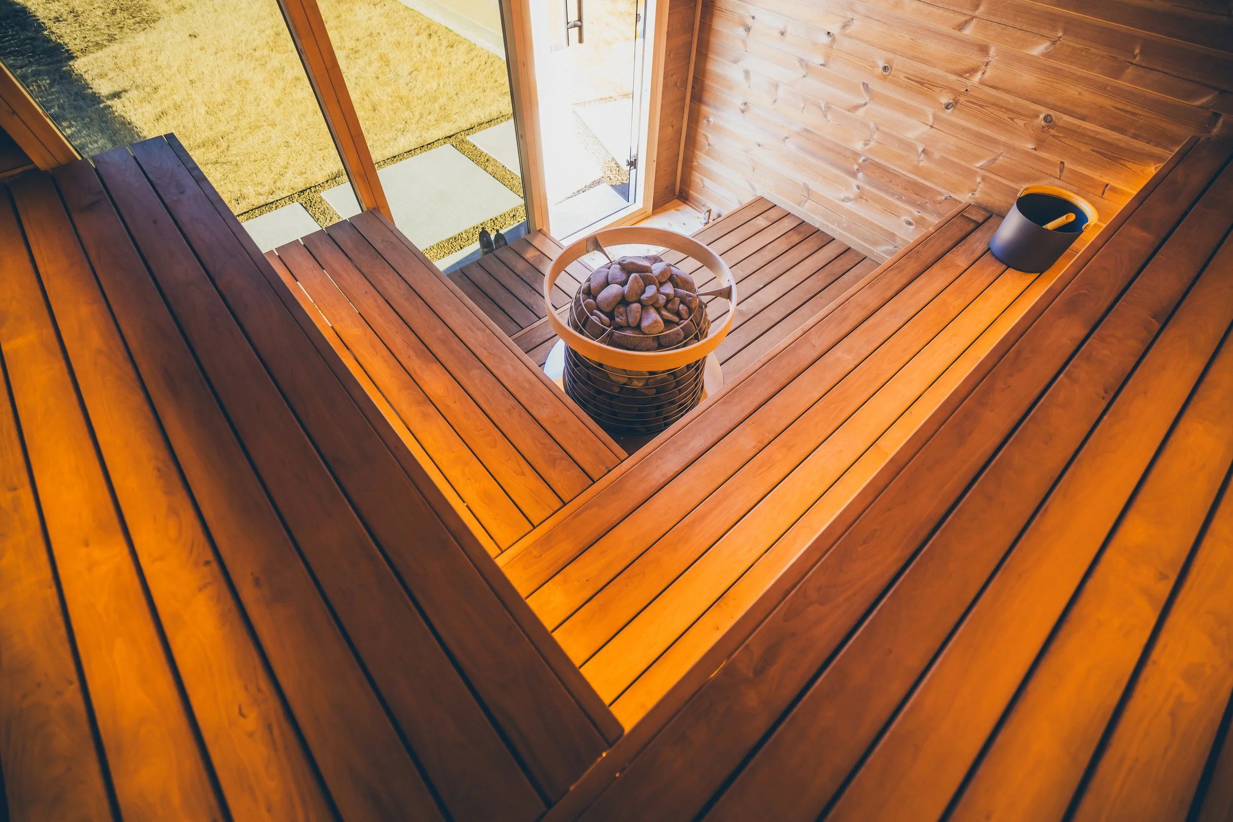 Interior of a wooden sauna with a stone-filled heater, bucket and ladle, and open door to a grassy yard.