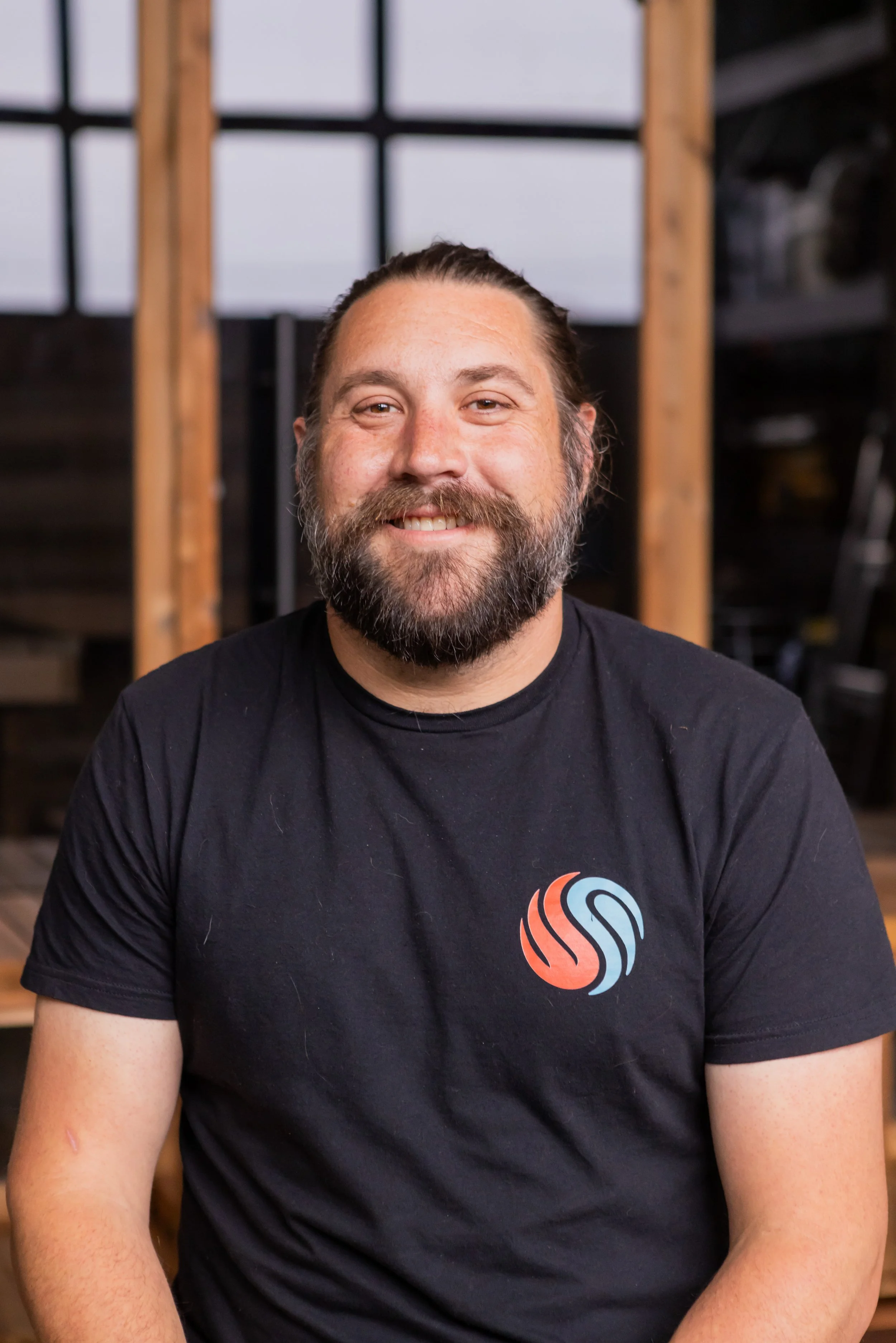 Chillfire Sauna Company team member sitting in front of a wooden sauna framework and windows, smiling and showcasing hands-on sauna craftsmanship.