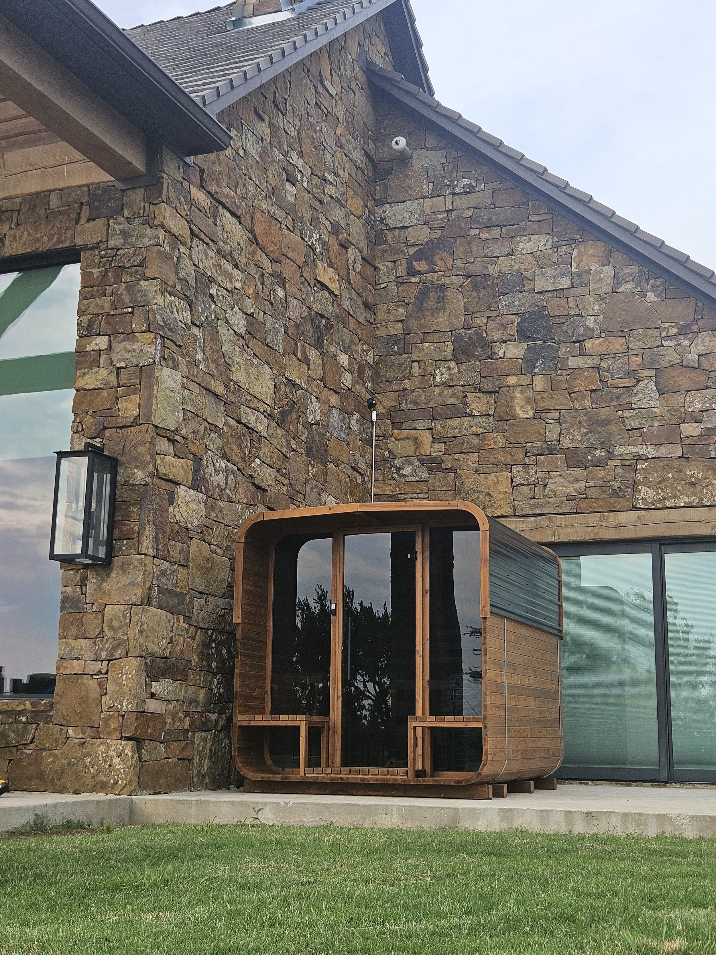 Small wooden outdoor sauna with curved roof on a concrete slab in front of a stone house.