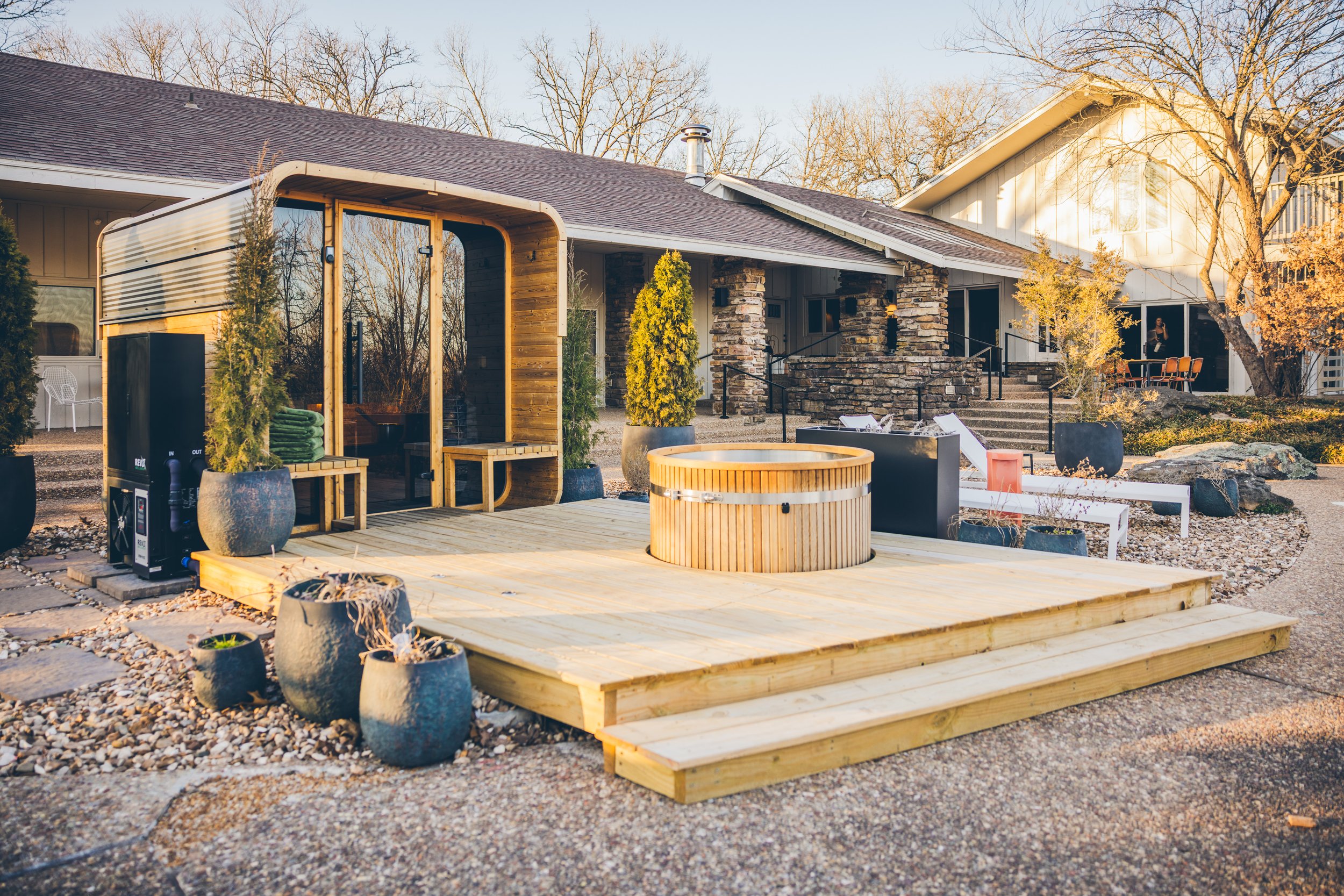 Exterior view of a lodge with a wooden deck, cold plunge tub and potted plants at sunset.