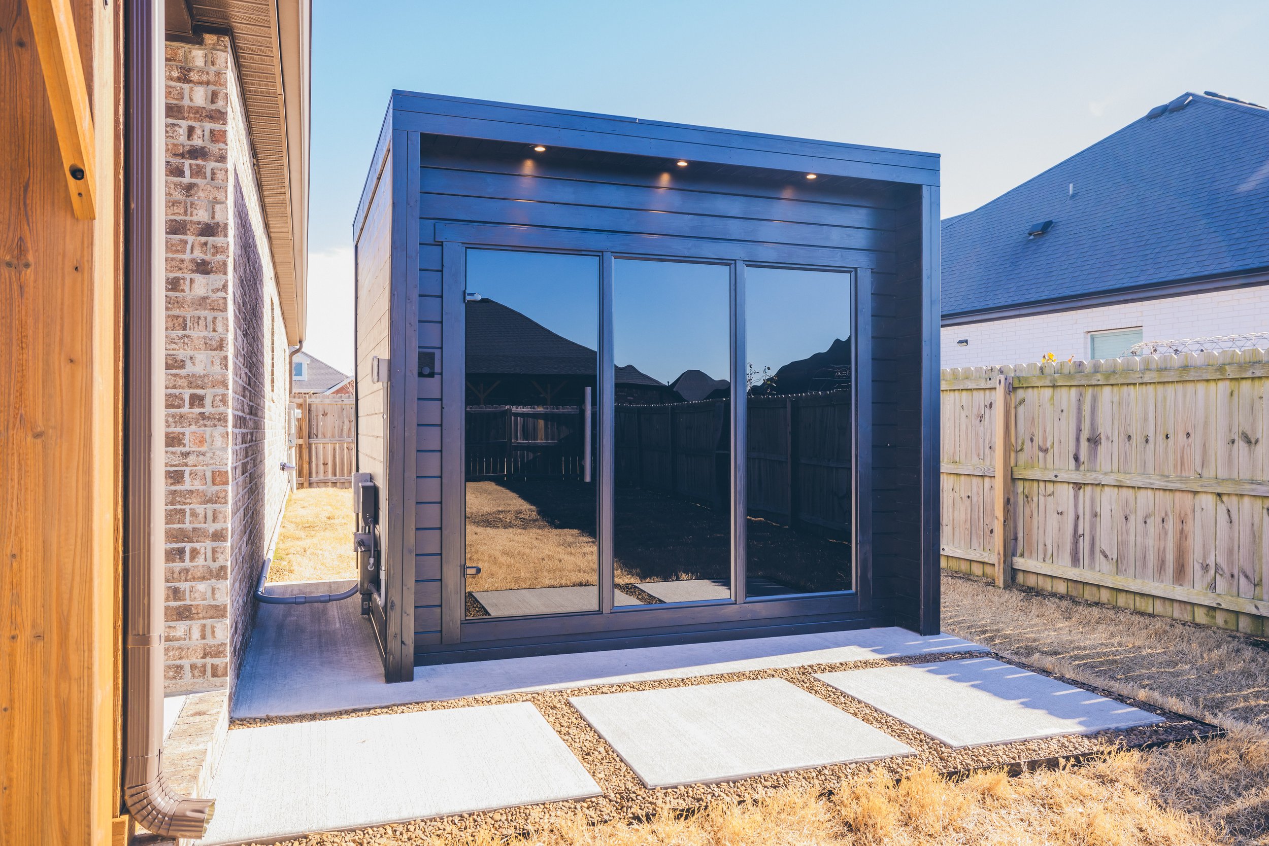 Modern backyard with a black wooden sauna cabin featuring glass double doors, next to a brick house.