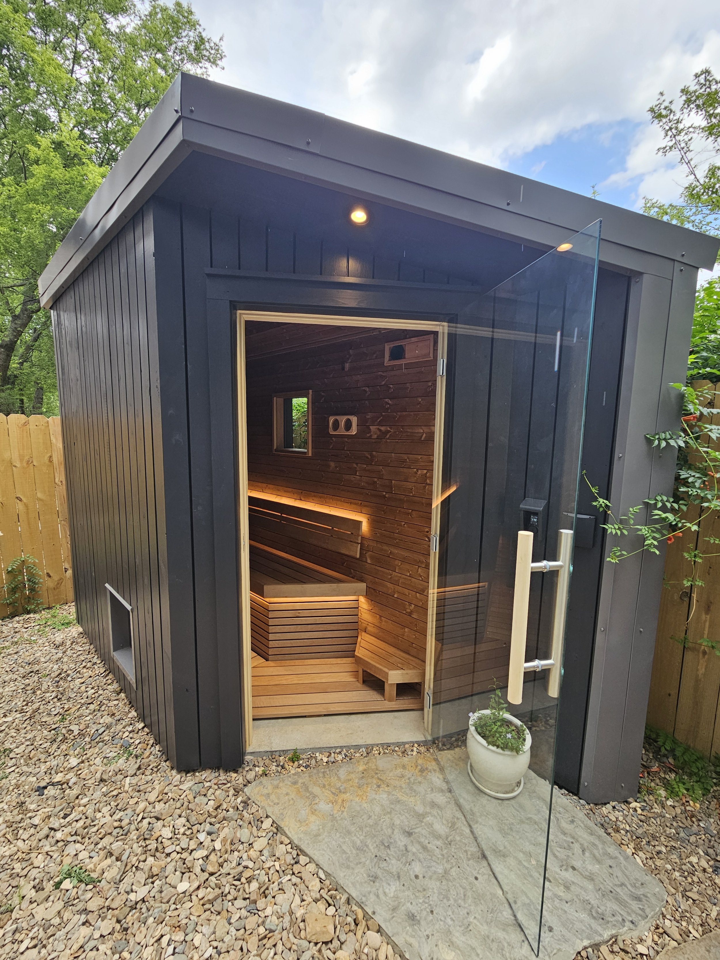 Small black wooden sauna with glass door and warm lighting, set outdoors on gravel.