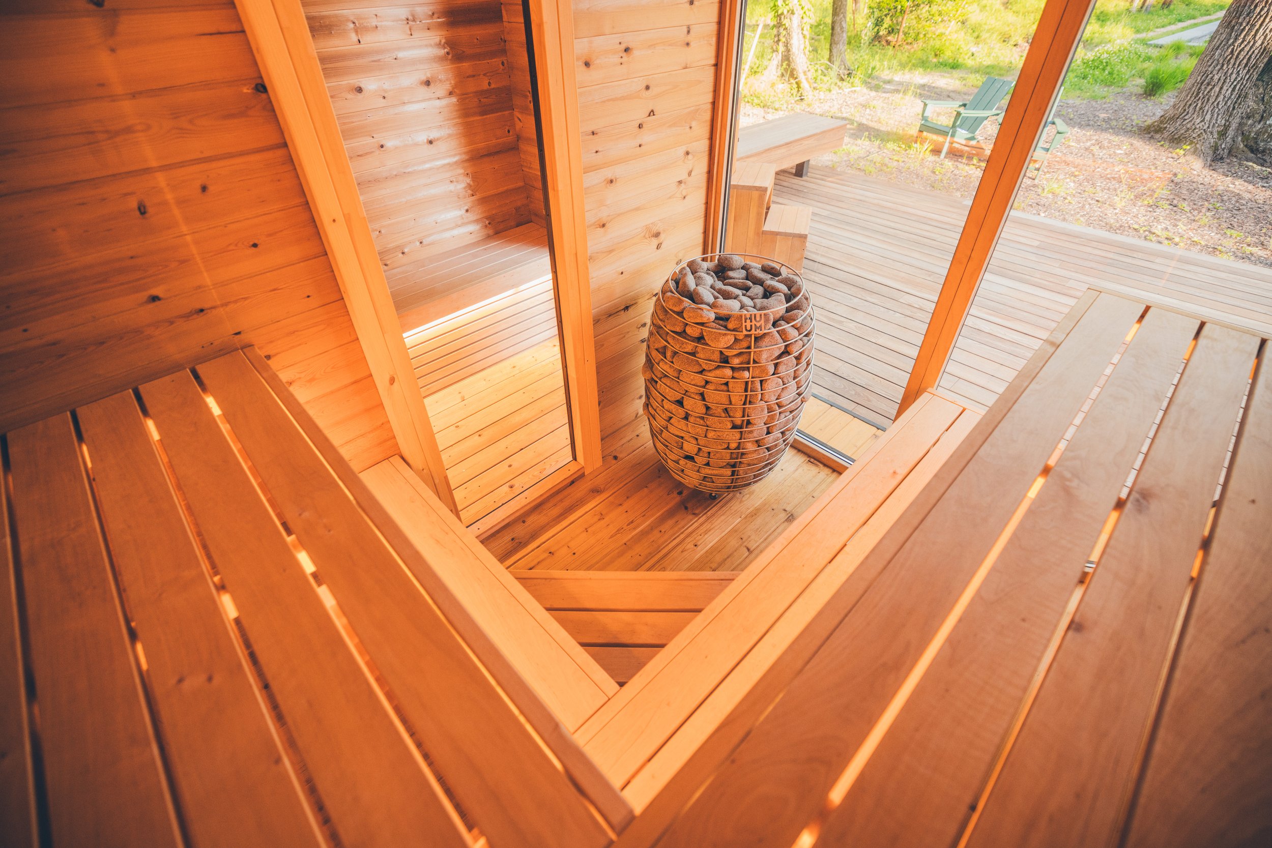 Interior of a wooden sauna with benches and a stone-filled heater, overlooking an outdoor deck.