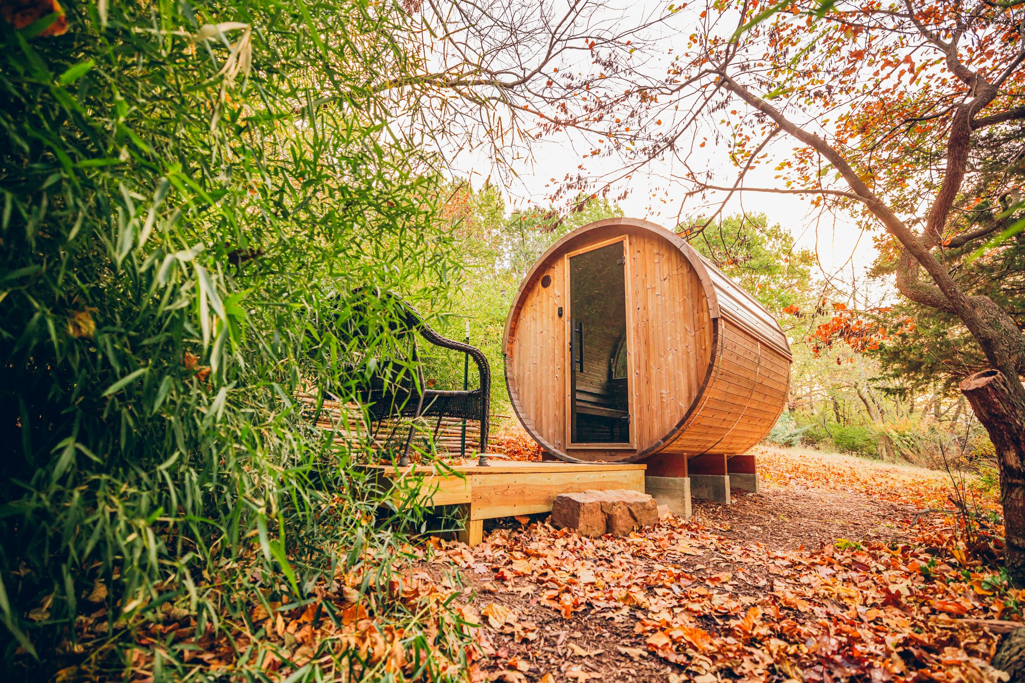 Round wooden barrel sauna with glass door elevated on piers, surrounded by autumn trees and fallen leaves.