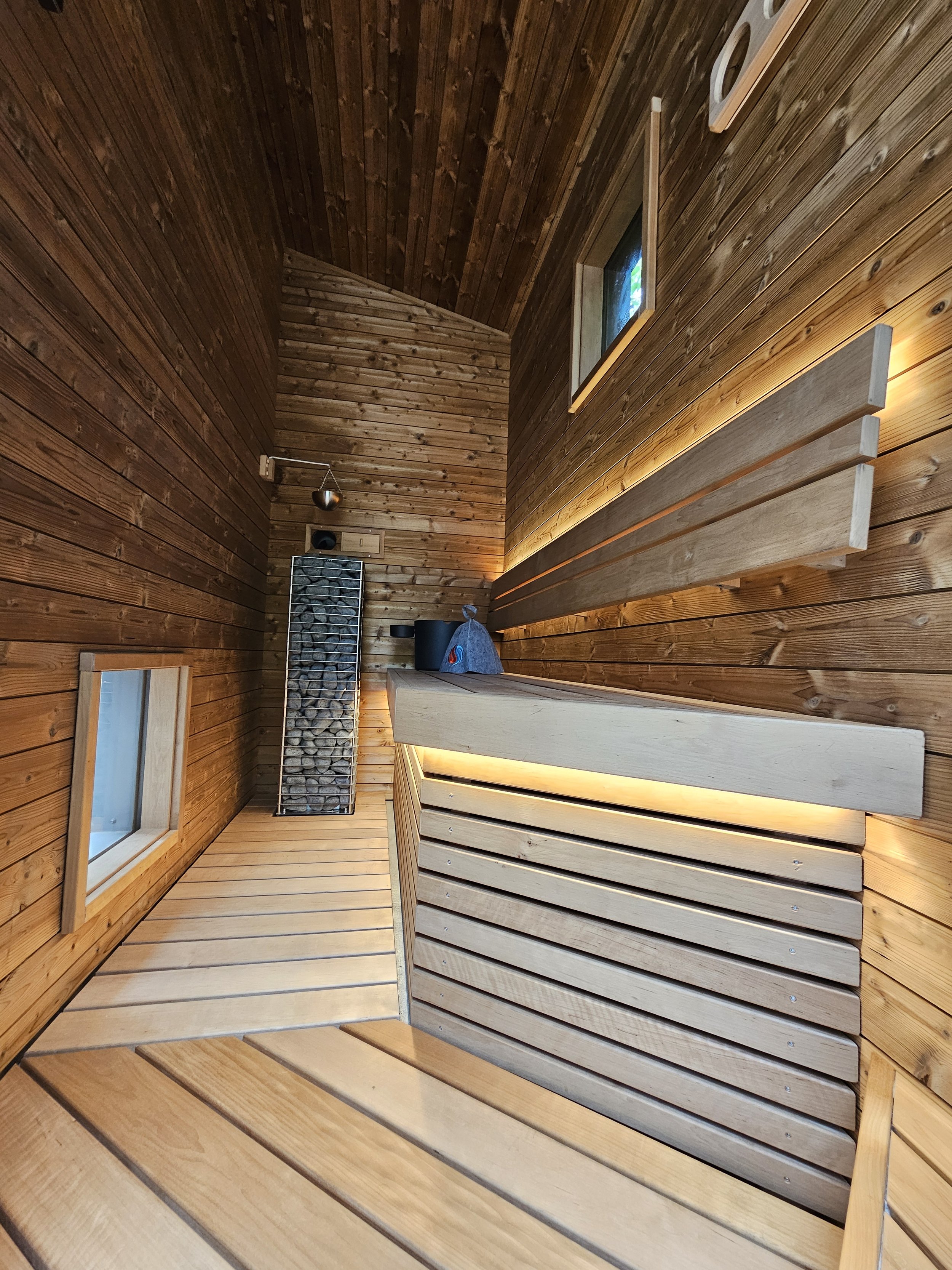 Interior of a small wooden sauna with benches, windows and a stone-filled heater.