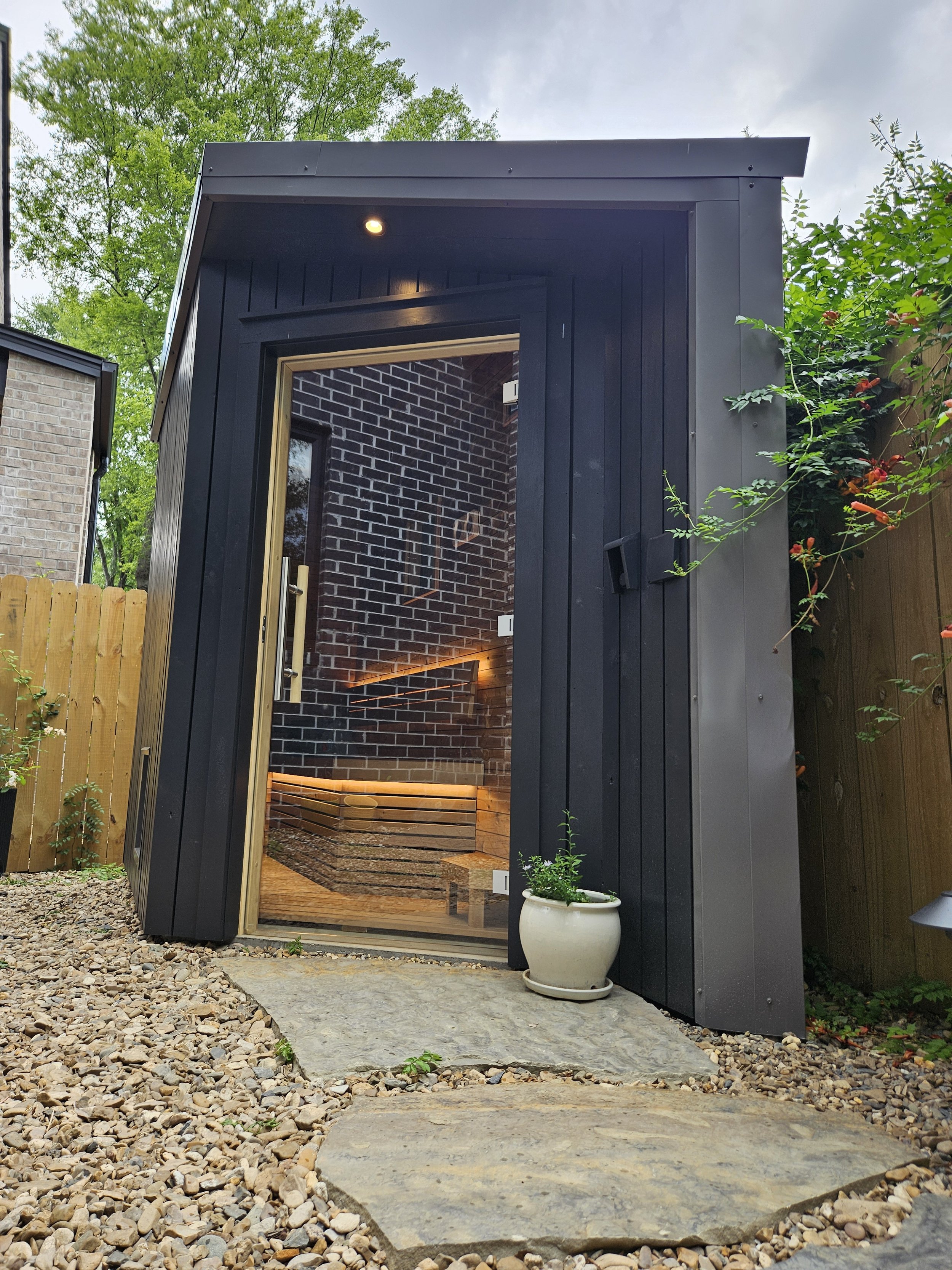 Small black wooden sauna with glass door on a stone path with plants, trees and a fence.