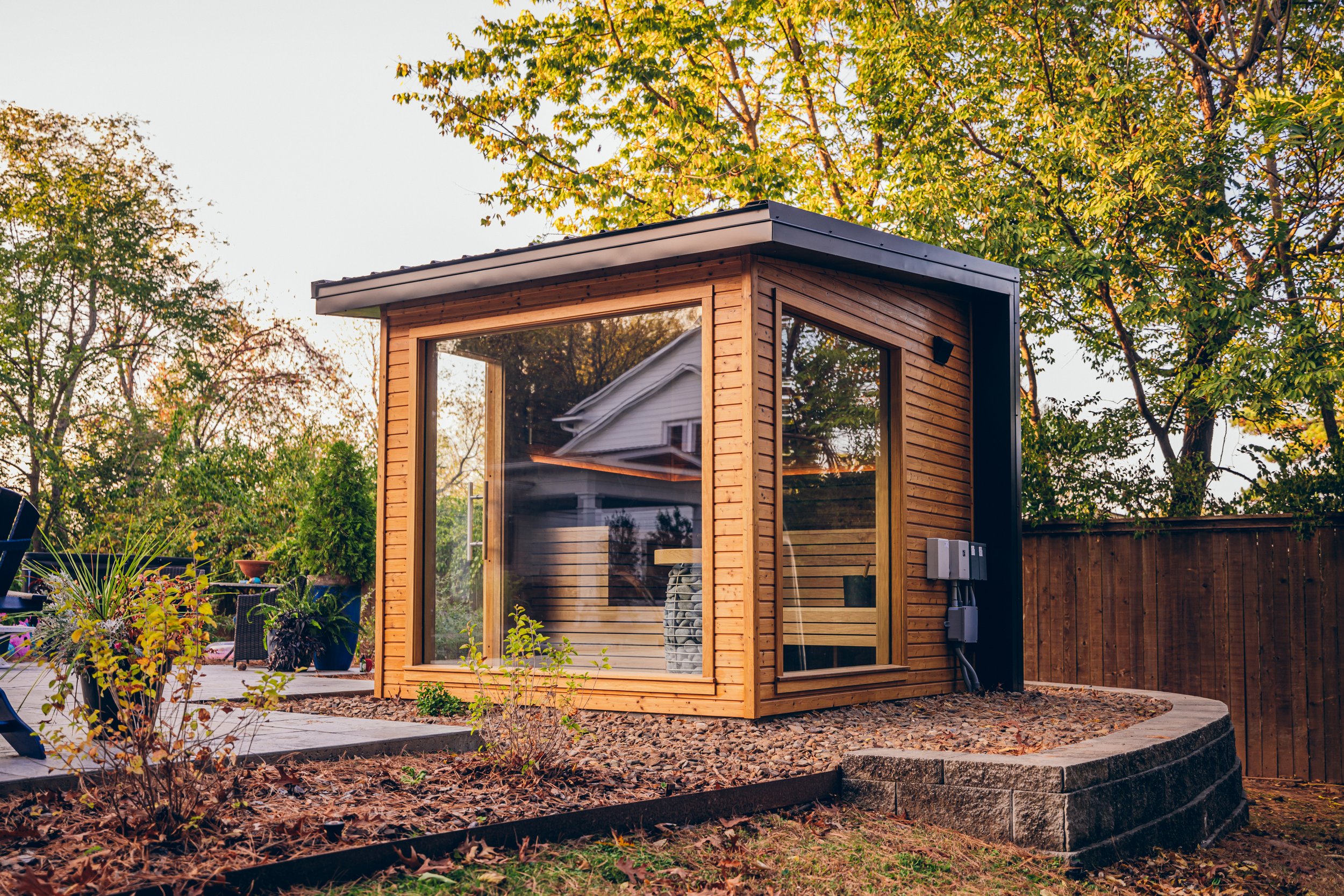 Small outdoor wooden sauna with large glass windows in a backyard with trees and a wooden fence.