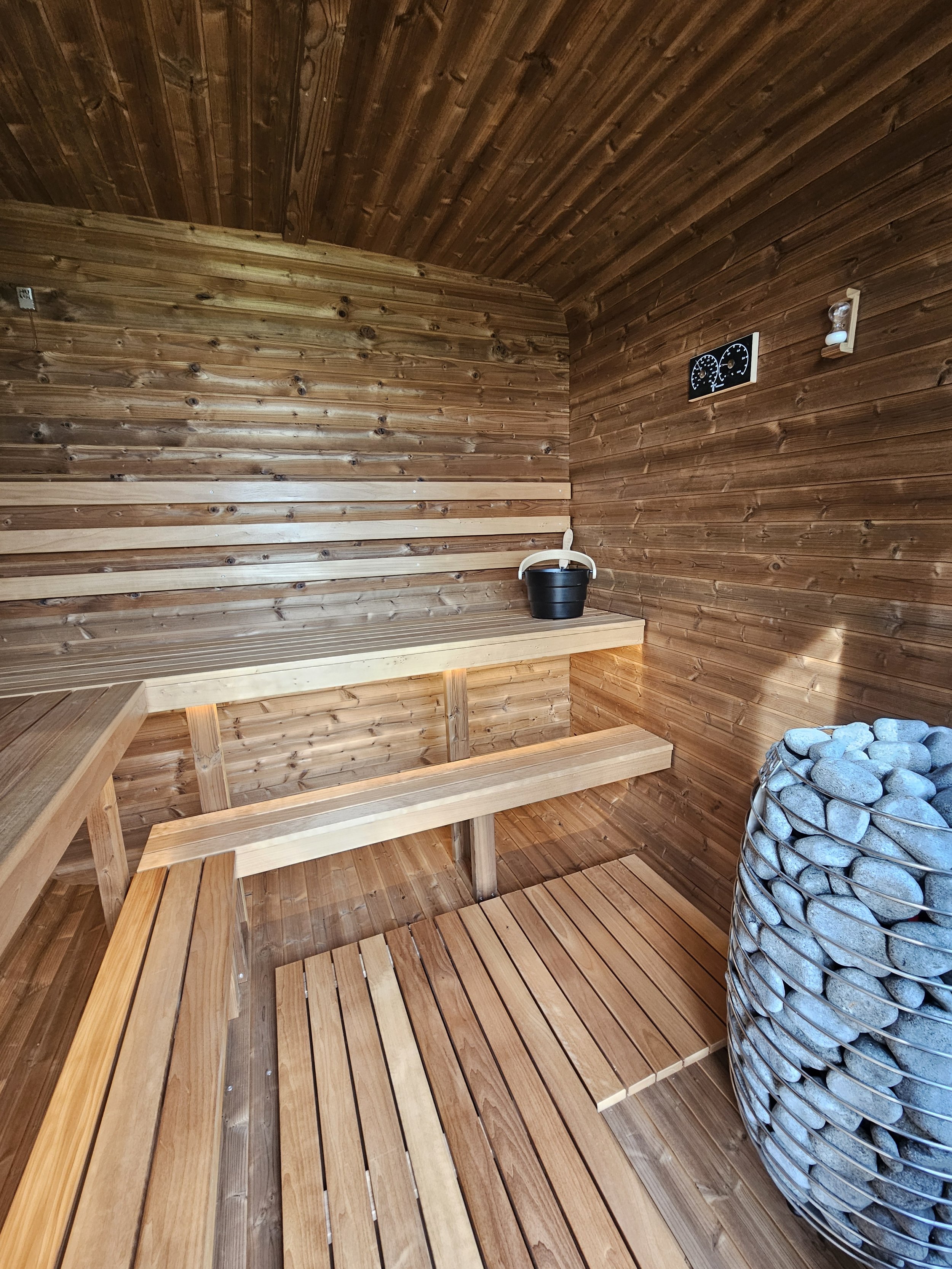 Wood-paneled sauna room with benches, bucket and ladle, heater with stones and a thermometer.