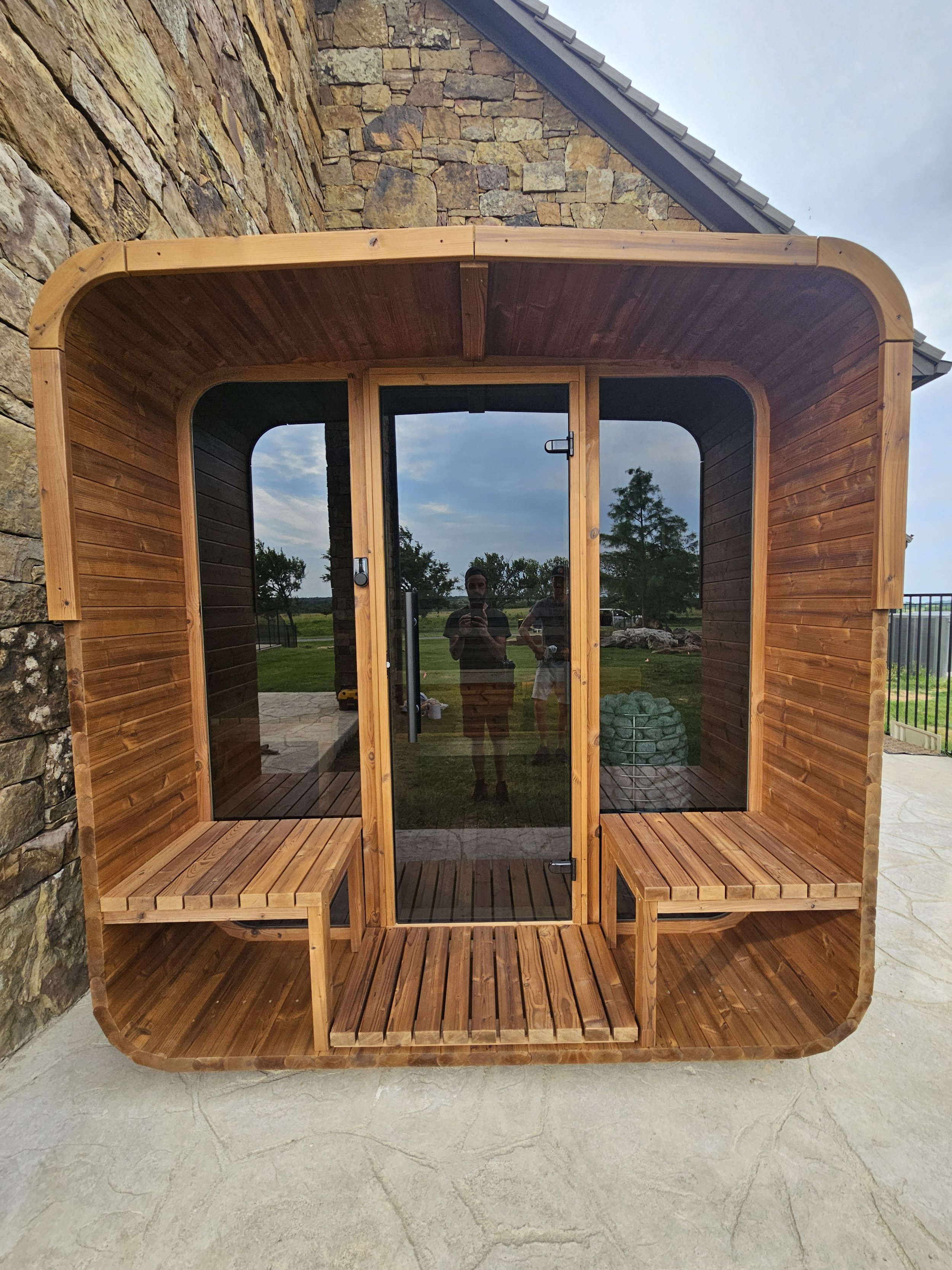 Small wooden sauna with glass door built against a stone wall, featuring two wooden benches.