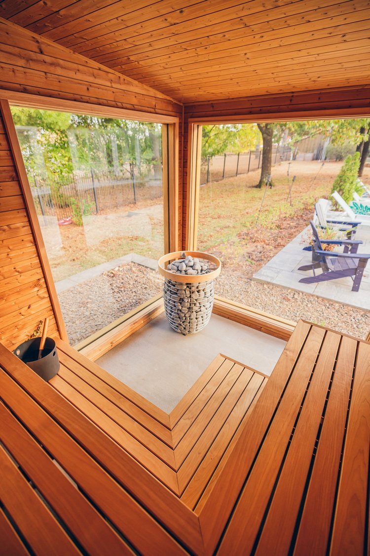 Interior of a sauna with wooden benches and a stone-filled heater, with large glass windows onto a backyard.