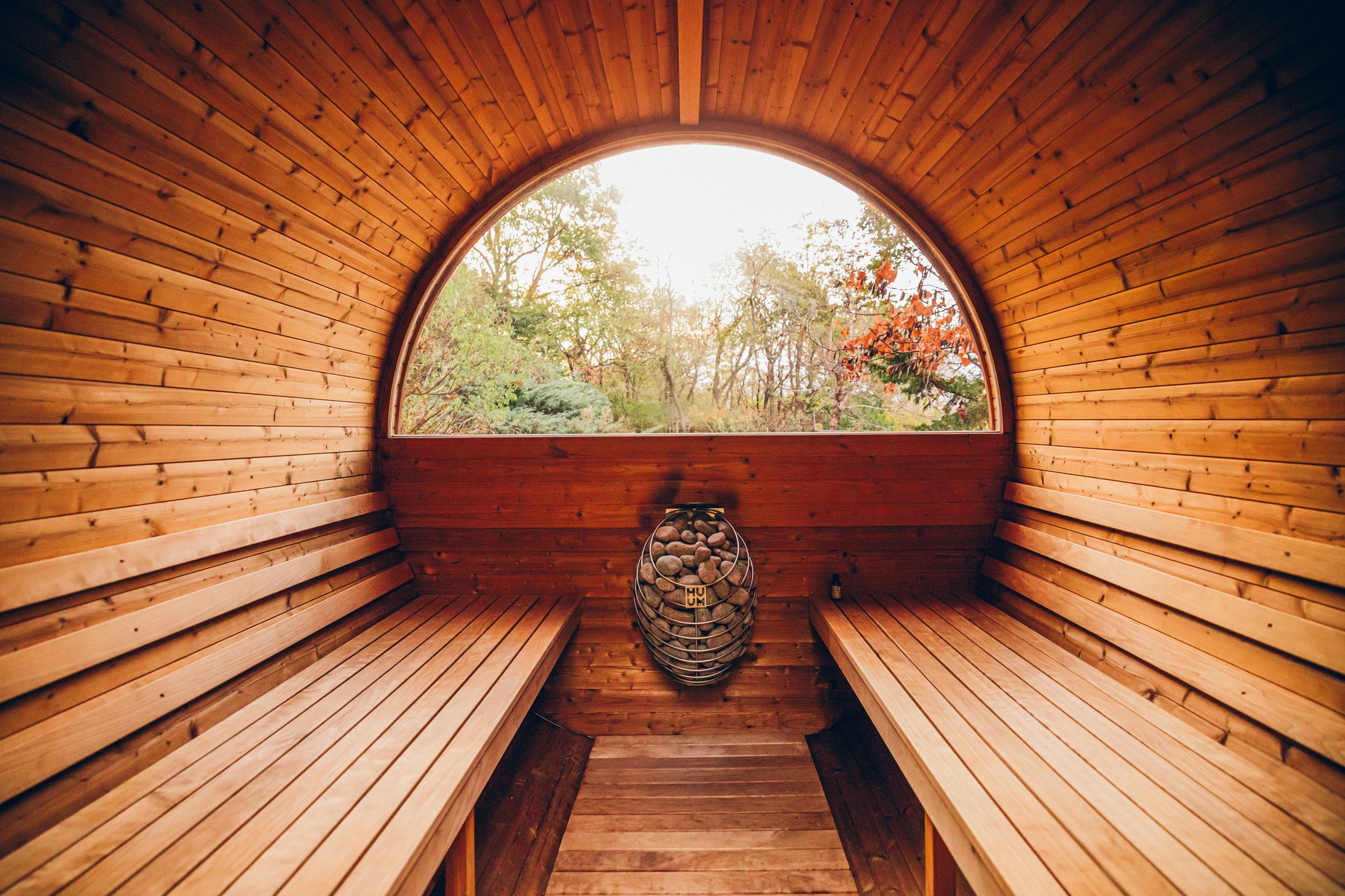 Interior of a barrel sauna with benches on both sides and a large circular window overlooking trees.